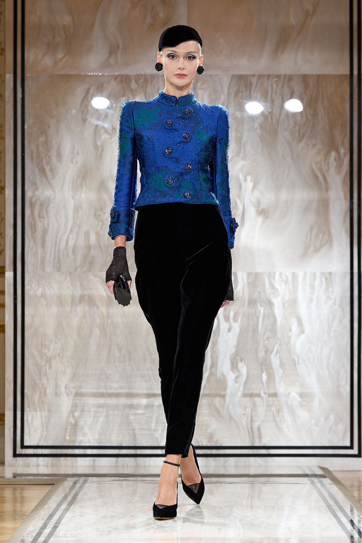 Model walking the runway in a blue embroidered jacket paired with black velvet pants and black accessories.