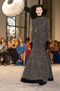 Model on a runway wearing a long black-and-white patterned gown with gloves; audience watches in the background.