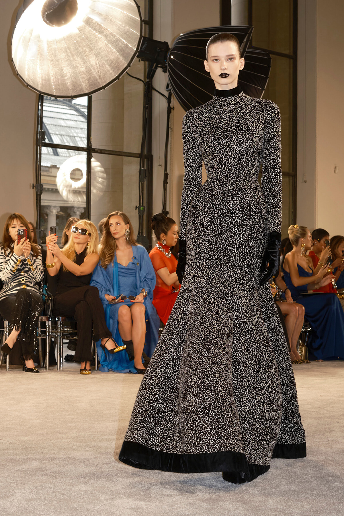 Model on a runway wearing a long black-and-white patterned gown with gloves; audience watches in the background.