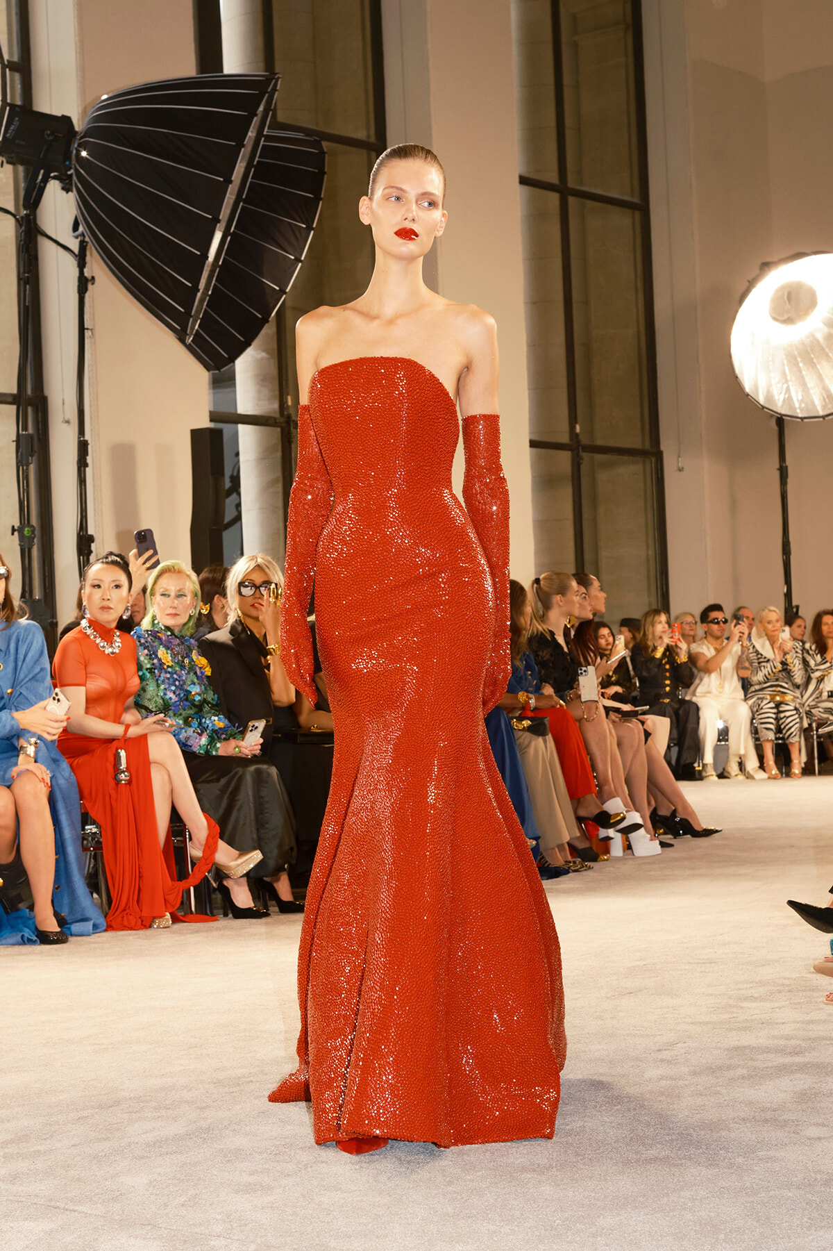 Model walking a runway in a strapless, floor-length red sequin gown with matching red gloves, audience watching.