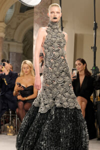 Model walks the runway in a high-neck, silver-black scalloped gown with a voluminous textured skirt; audience members photograph the moment in the background.