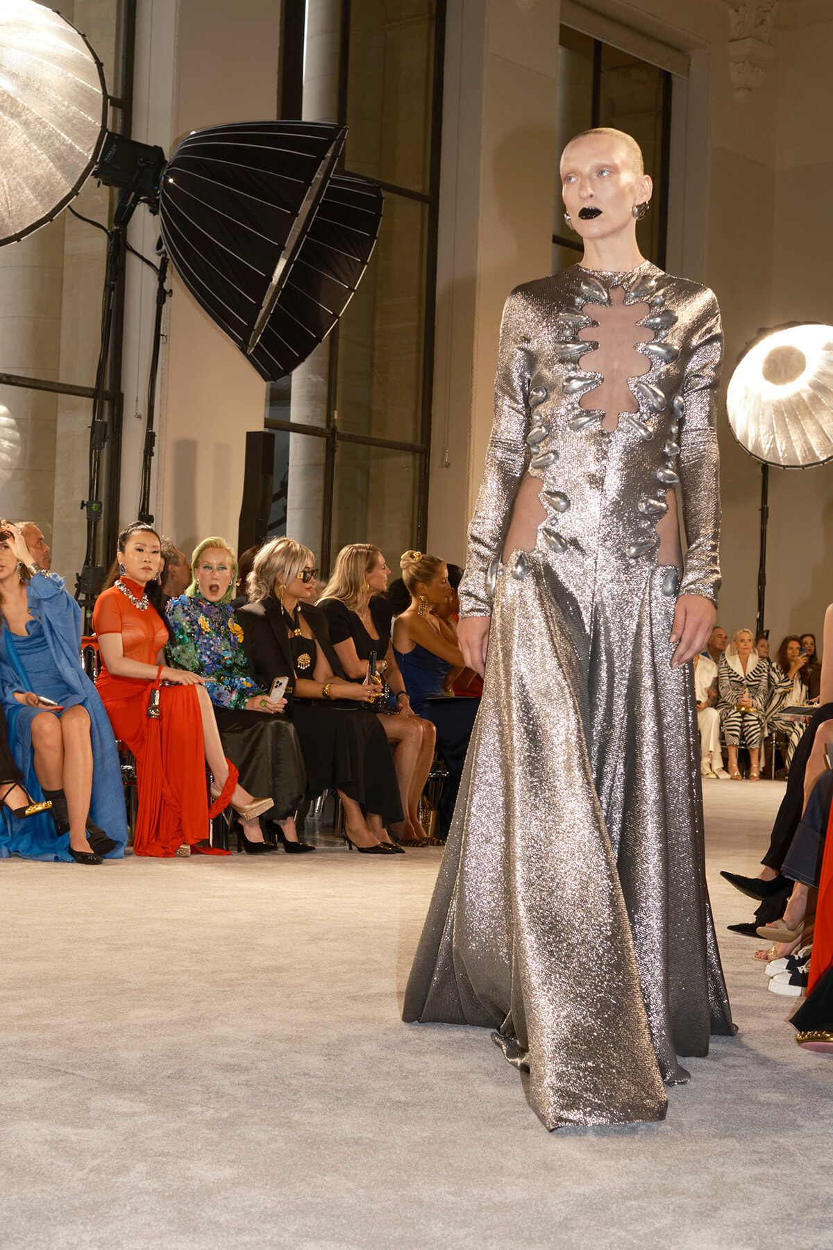 Model walks the runway in a shimmering silver gown with sculpted cutouts, audience seated along the catwalk behind her.