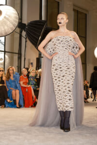 Model on a runway wearing a silver beaded strapless dress with an oval eye-pattern, paired with a sheer cape and dark lipstick, posing with hands on hips as photographers watch from the audience.