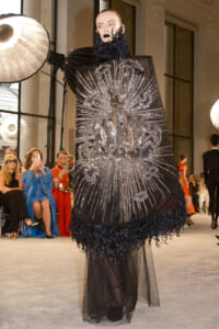 Model on a runway wearing a dramatic black cape with a radiating silver beaded design and sheer hem, surrounded by seated fashion show guests and photographers.