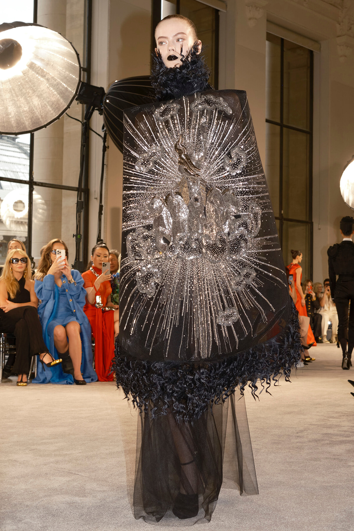 Model on a runway wearing a dramatic black cape with a radiating silver beaded design and sheer hem, surrounded by seated fashion show guests and photographers.