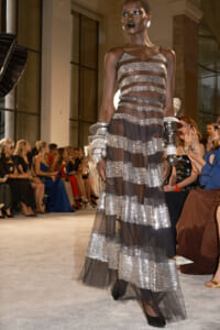 Model walking a fashion runway in a sheer, striped metallic gown with pronounced silver bands and black underlay, wearing bold jewelry and dark lipstick.