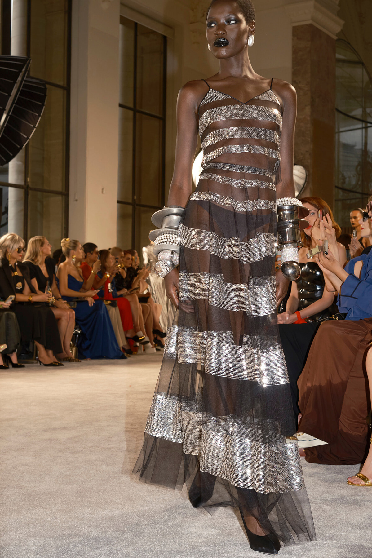Model walking a fashion runway in a sheer, striped metallic gown with pronounced silver bands and black underlay, wearing bold jewelry and dark lipstick.