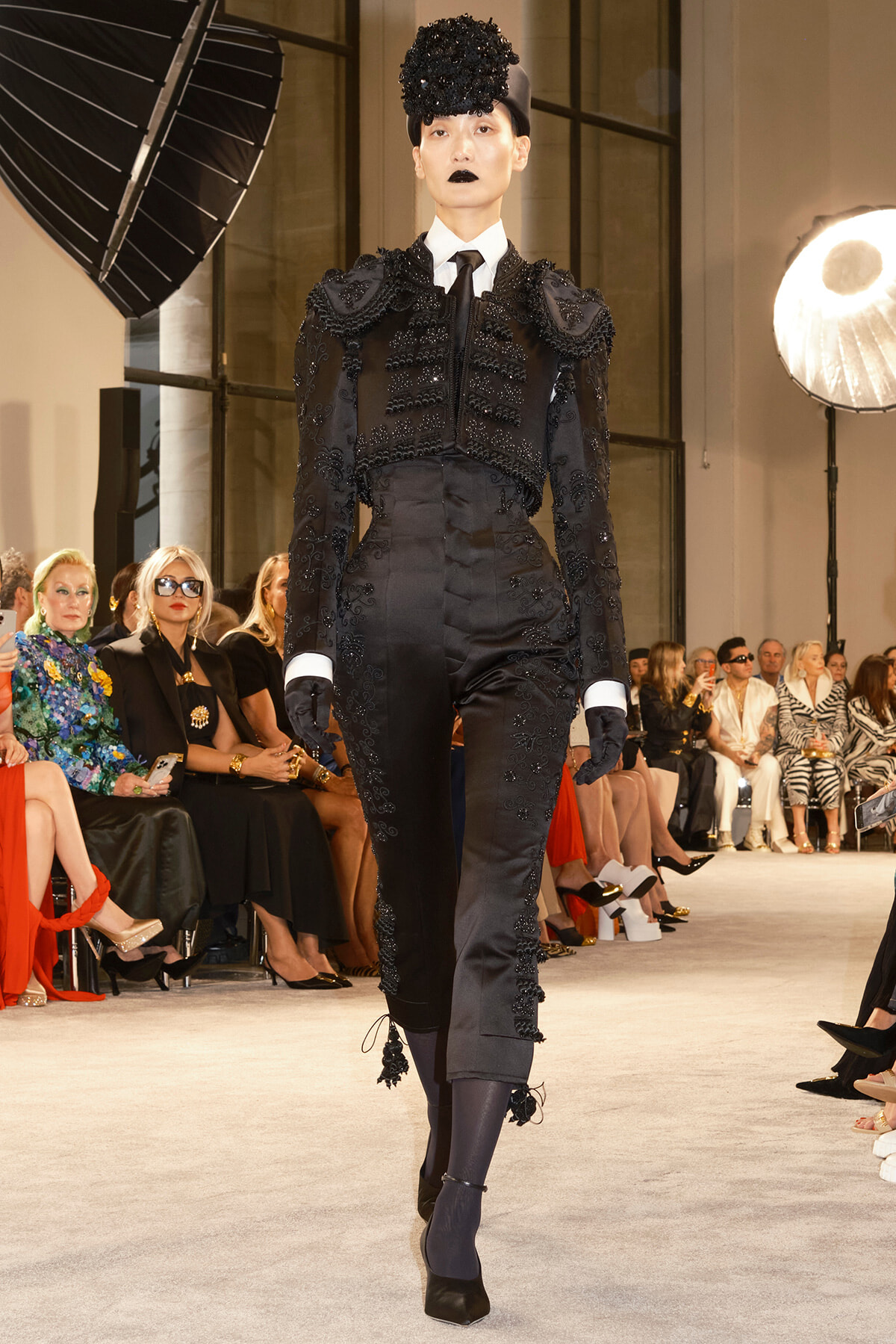 Model walks the runway in a gothic, black embellished suit with gloves and a tall hat, audience watching in the background.