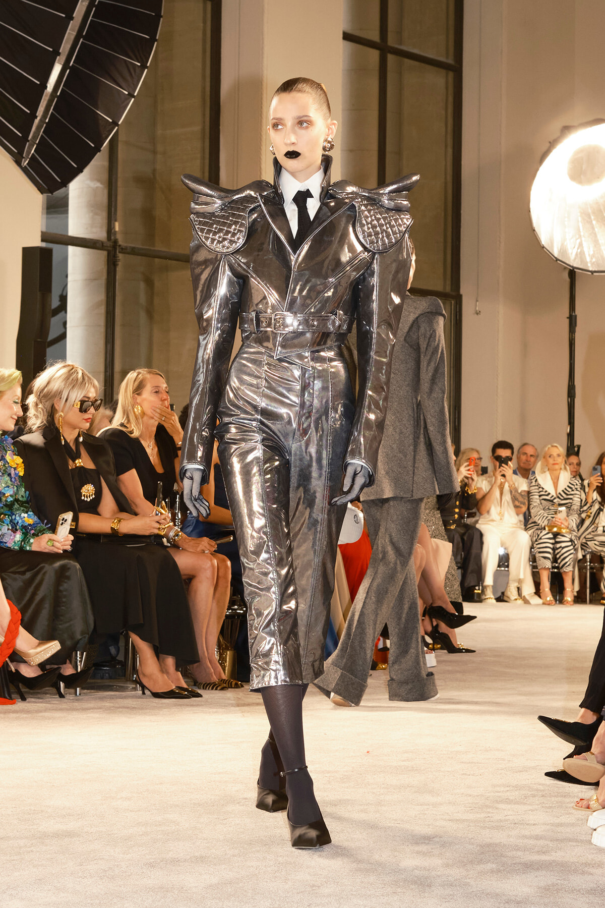 Model on a runway wearing a metallic silver armored-style coat with oversized shoulders and a belt.