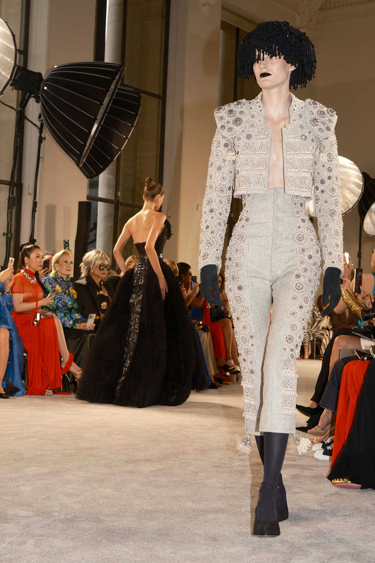 Model walks the runway in a silver embellished suit with bold shoulder details and navy gloves.