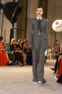 Model walks the runway in a gray textured suit with oversized shoulders and a black polka-dot bow, audience seated along the catwalk.