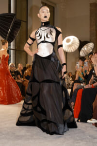 Model walks a fashion runway wearing a black-and-white avant-garde gown with a sheer layered skirt and statement jewelry, audience watching.