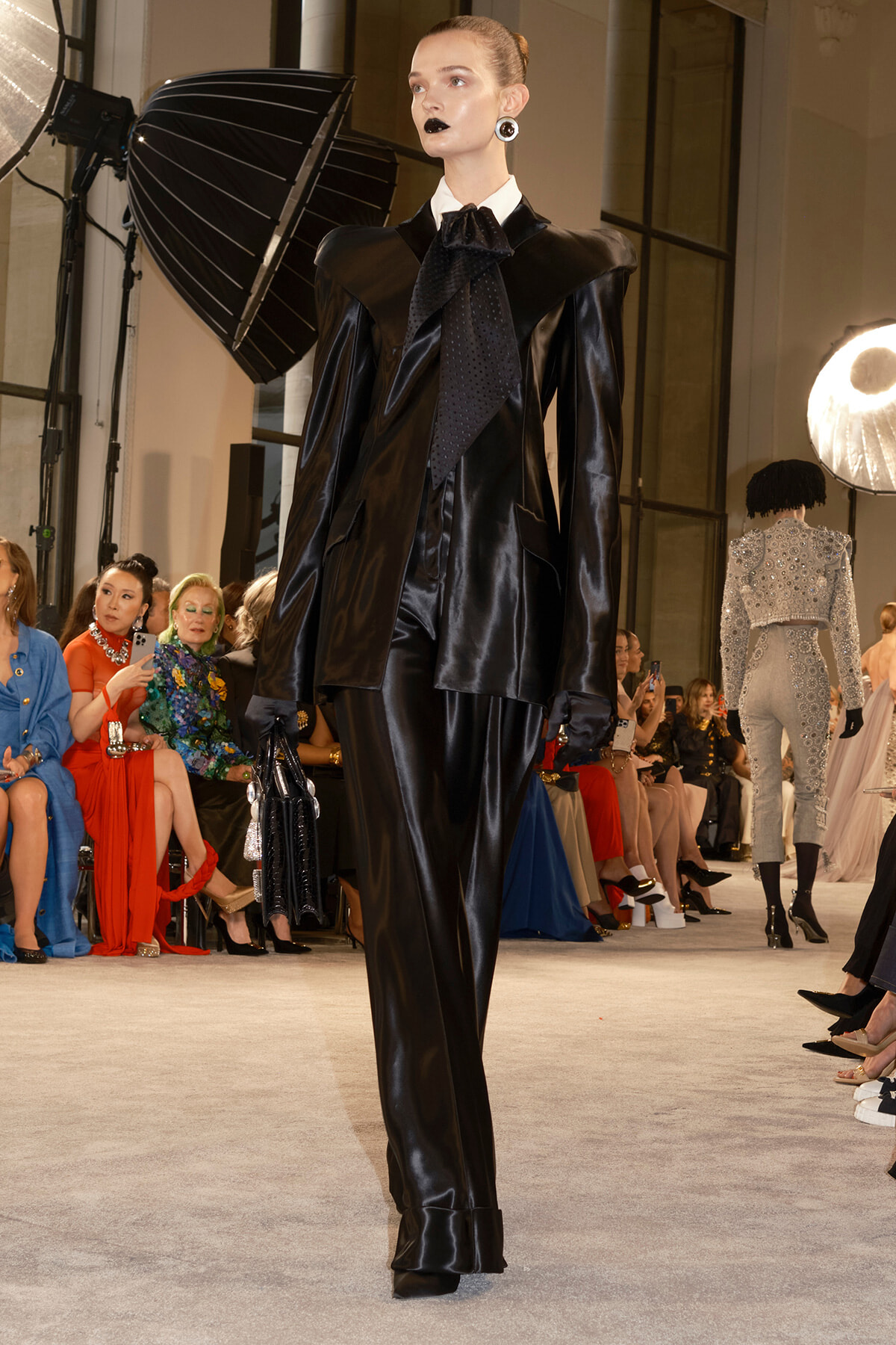 Model walks the runway in a glossy black patent-leather suit with oversized shoulder details and a large bow-tie, holding a black handbag.