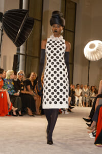 Model on a runway wearing a white sleeveless polka-dot dress, navy gloves, and a large pearl necklace with a tall black headwrap.