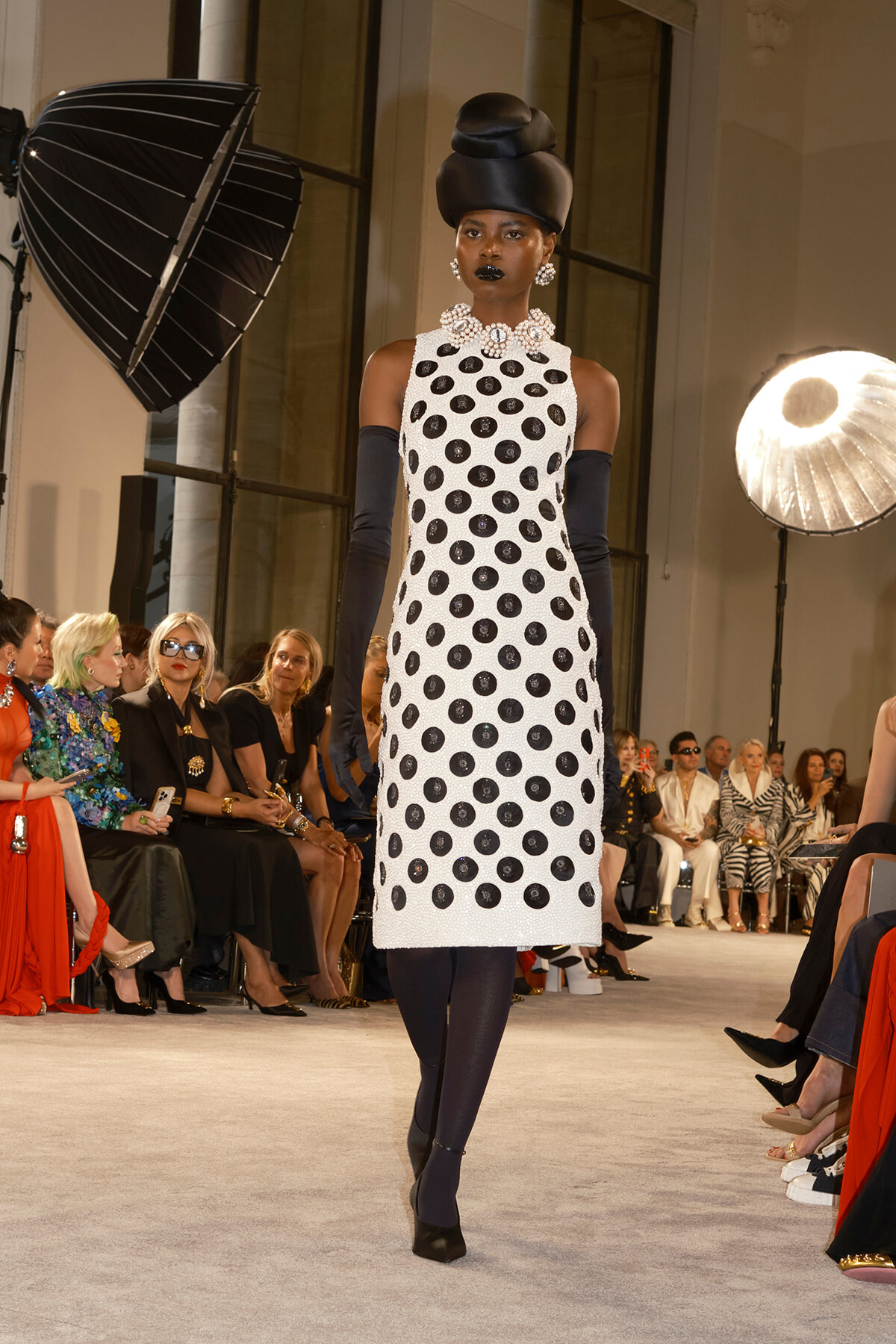 Model on a runway wearing a white sleeveless polka-dot dress, navy gloves, and a large pearl necklace with a tall black headwrap.