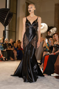Model on a runway wearing a black satin gown with sheer beaded panels and a mermaid train.