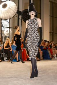 Model on runway wearing a sleeveless silver beaded dress with large circular embellishments, paired with black elbow-long gloves and a tall textured black hat.