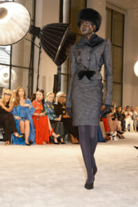 Model struts a gray tailored suit with fur collar and hat on a runway, audience watching in chic attire behind.