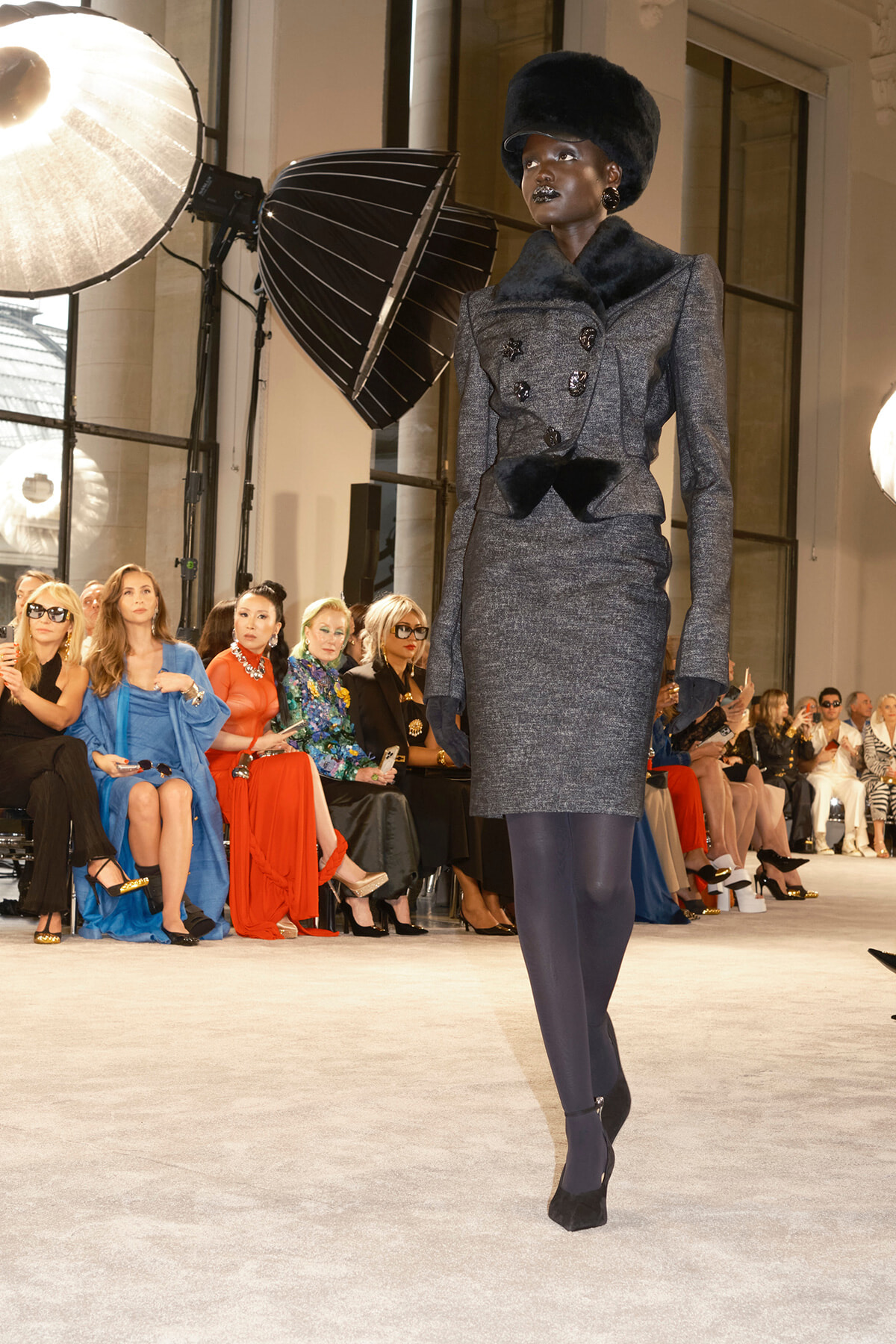 Model struts a gray tailored suit with fur collar and hat on a runway, audience watching in chic attire behind.