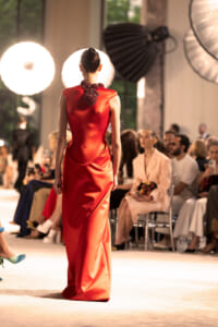 Back view of a model in a red satin gown with a sculpted beaded collar walking the runway, audience seated along the catwalk in soft focus.