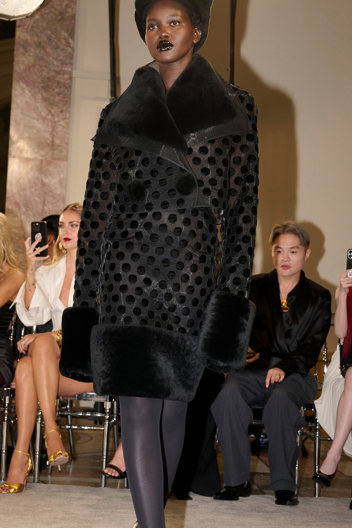 Model walks a runway in a black polka-dot coat with oversized fur collar and cuffs; audience seated in background.