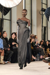 Model on a runway wearing a metallic gray off-the-shoulder gown with long matching gloves, mid-stride as spectators watch from the sides.