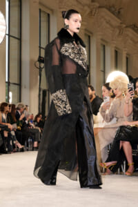 Model walks the runway in a sheer black coat with gold leaf embroidery and a black fur collar; audience sits along the catwalk.