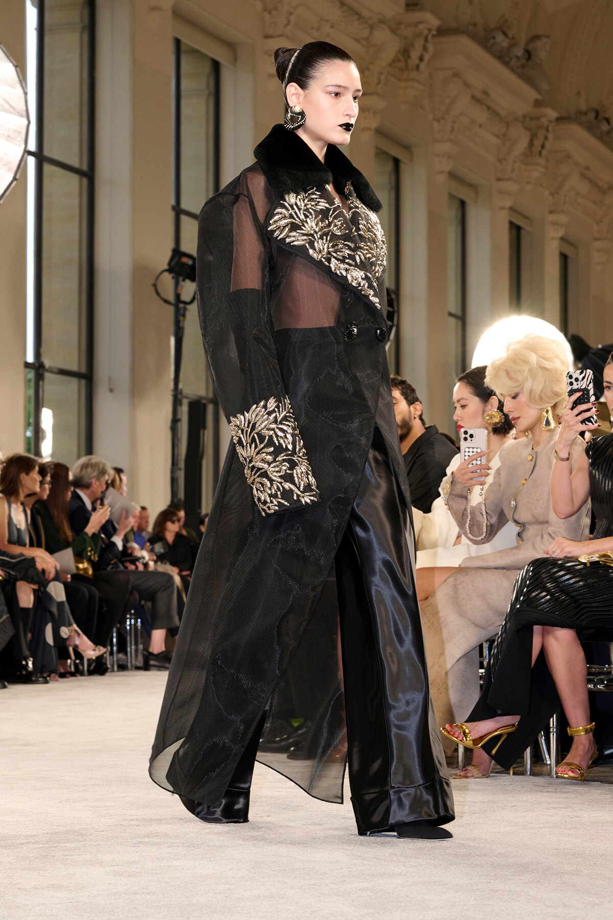 Model walks the runway in a sheer black coat with gold leaf embroidery and a black fur collar; audience sits along the catwalk.