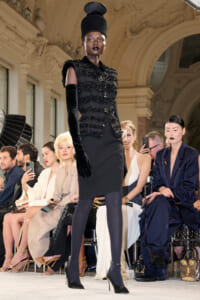 Model in a black beaded jacket, tall hat, and long velvet gloves walks a fashion-show runway as seated spectators watch.