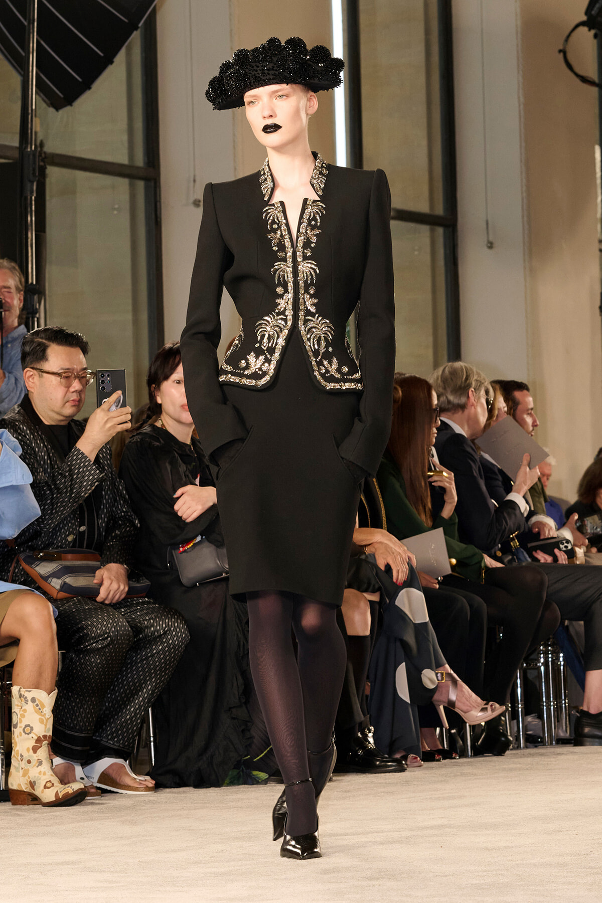 Model on a runway wearing a black tailored dress with silver embroidered detailing and a sculptural black hat with bold makeup, front view