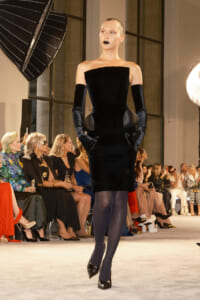 Model walks a runway in a strapless black velvet dress with long matching gloves and black heels, audience watching from the sides.
