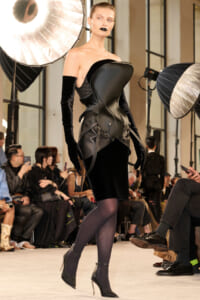 Model walking a fashion runway in a sculptural black leather corset dress with long velvet gloves and black heels, audience watching in the background