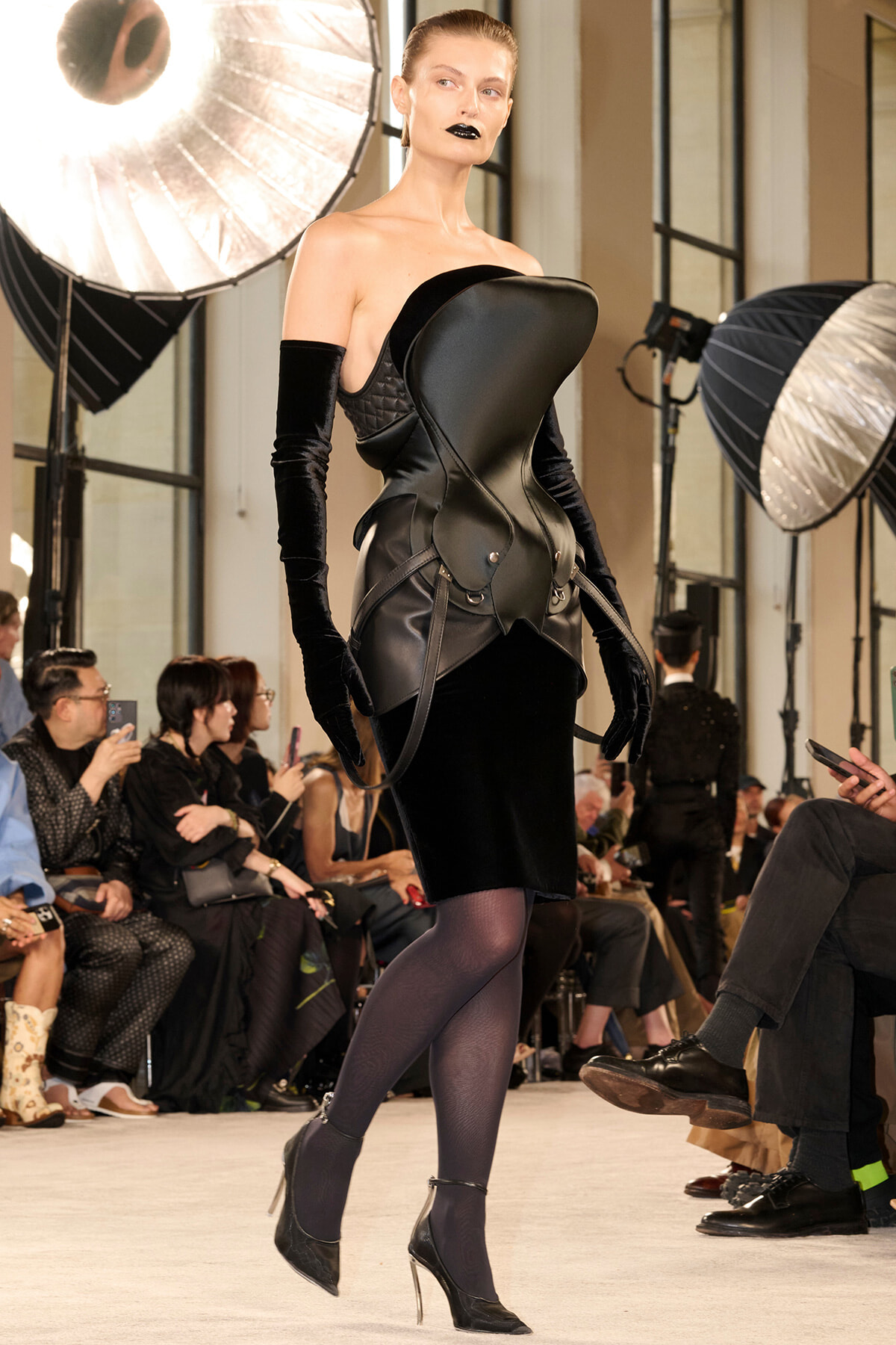 Model walking a fashion runway in a sculptural black leather corset dress with long velvet gloves and black heels, audience watching in the background