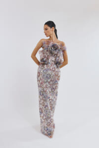 Full-length floral lace gown with large 3D flower appliqués at the bodice, worn by a model posing with hands on hips in profile.