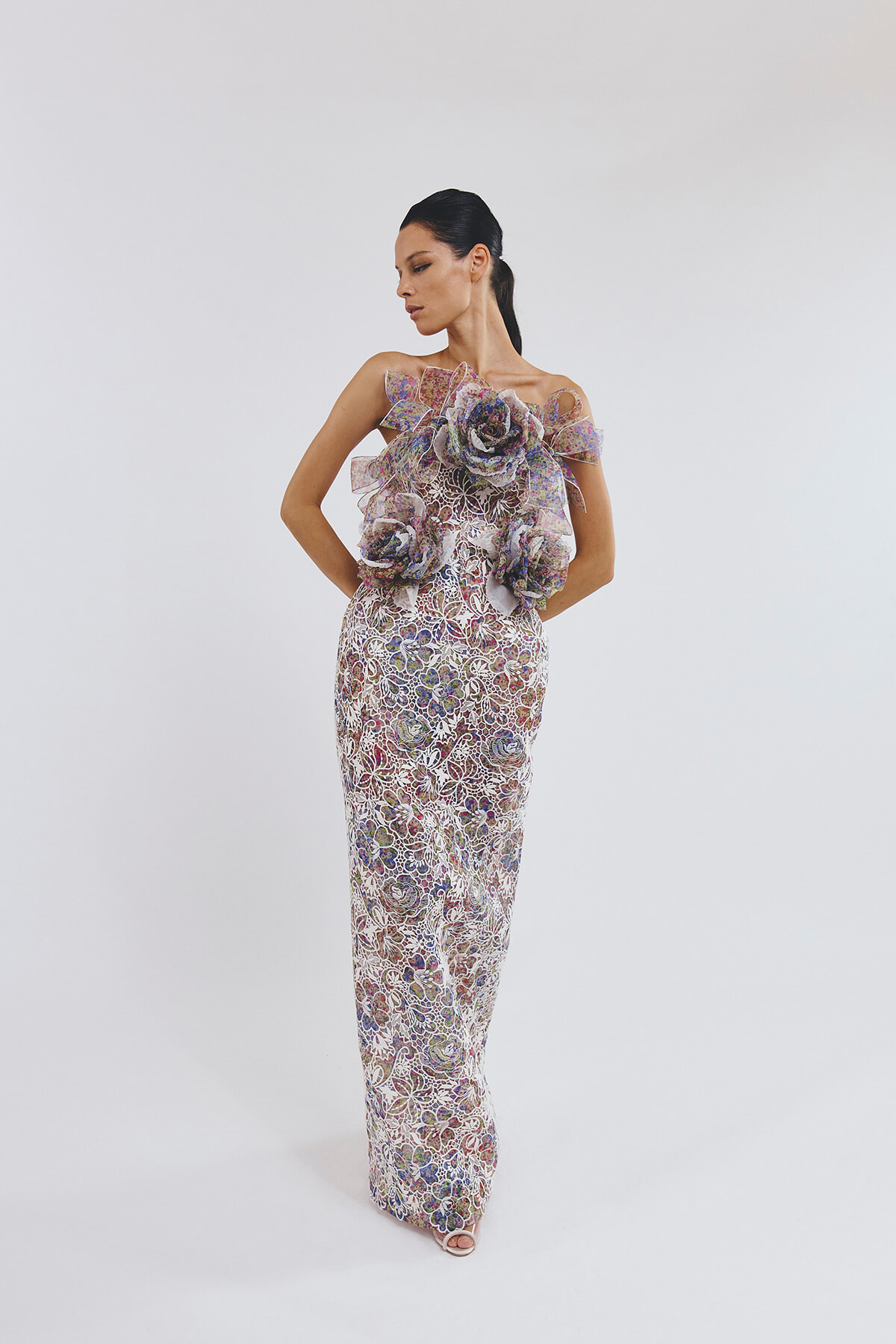 Full-length floral lace gown with large 3D flower appliqués at the bodice, worn by a model posing with hands on hips in profile.