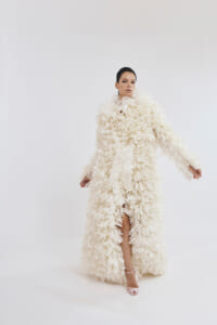 Model wearing an extravagant white feathered gown with high collar, posing against a light backdrop, left leg visible through a slit