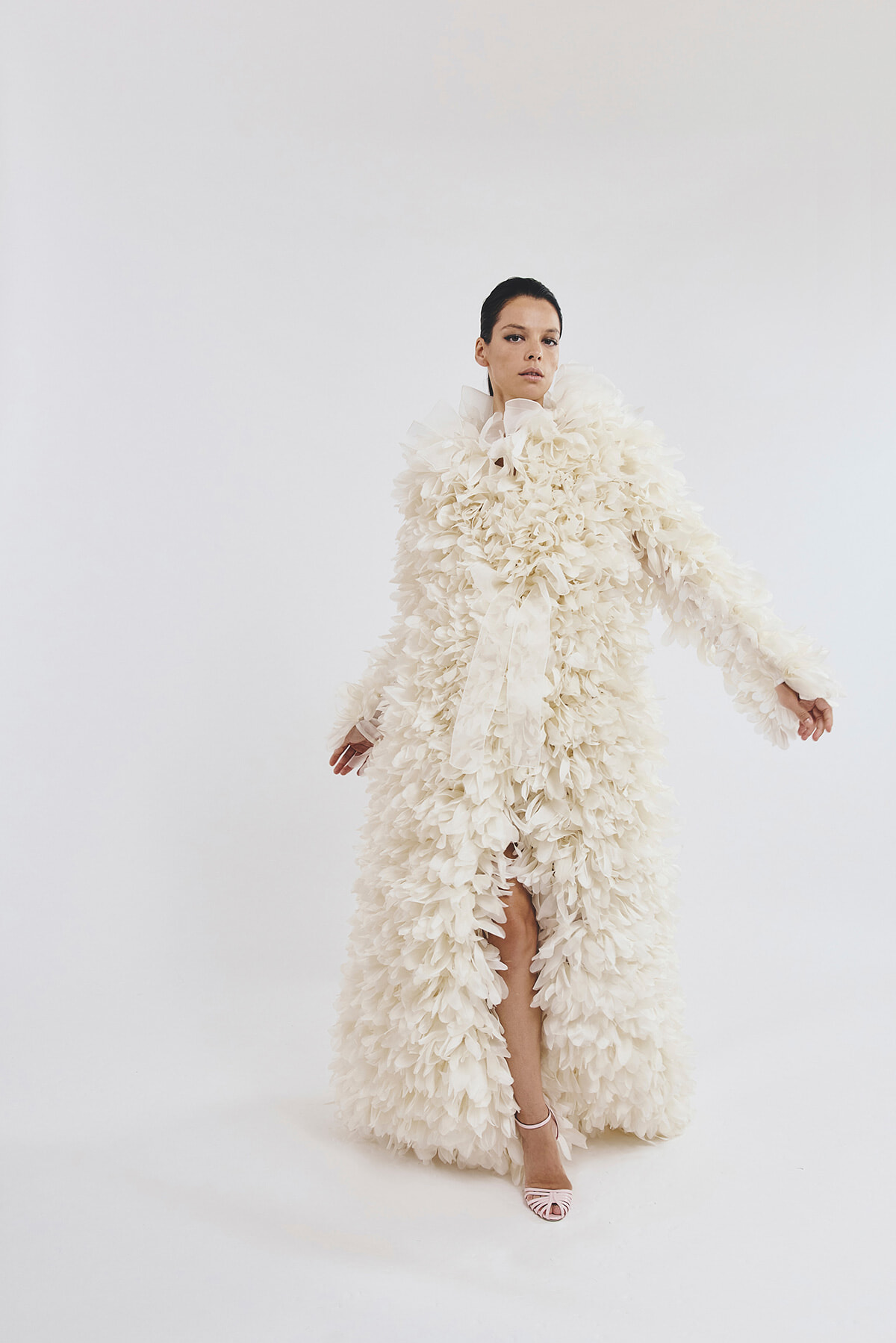 Model wearing an extravagant white feathered gown with high collar, posing against a light backdrop, left leg visible through a slit