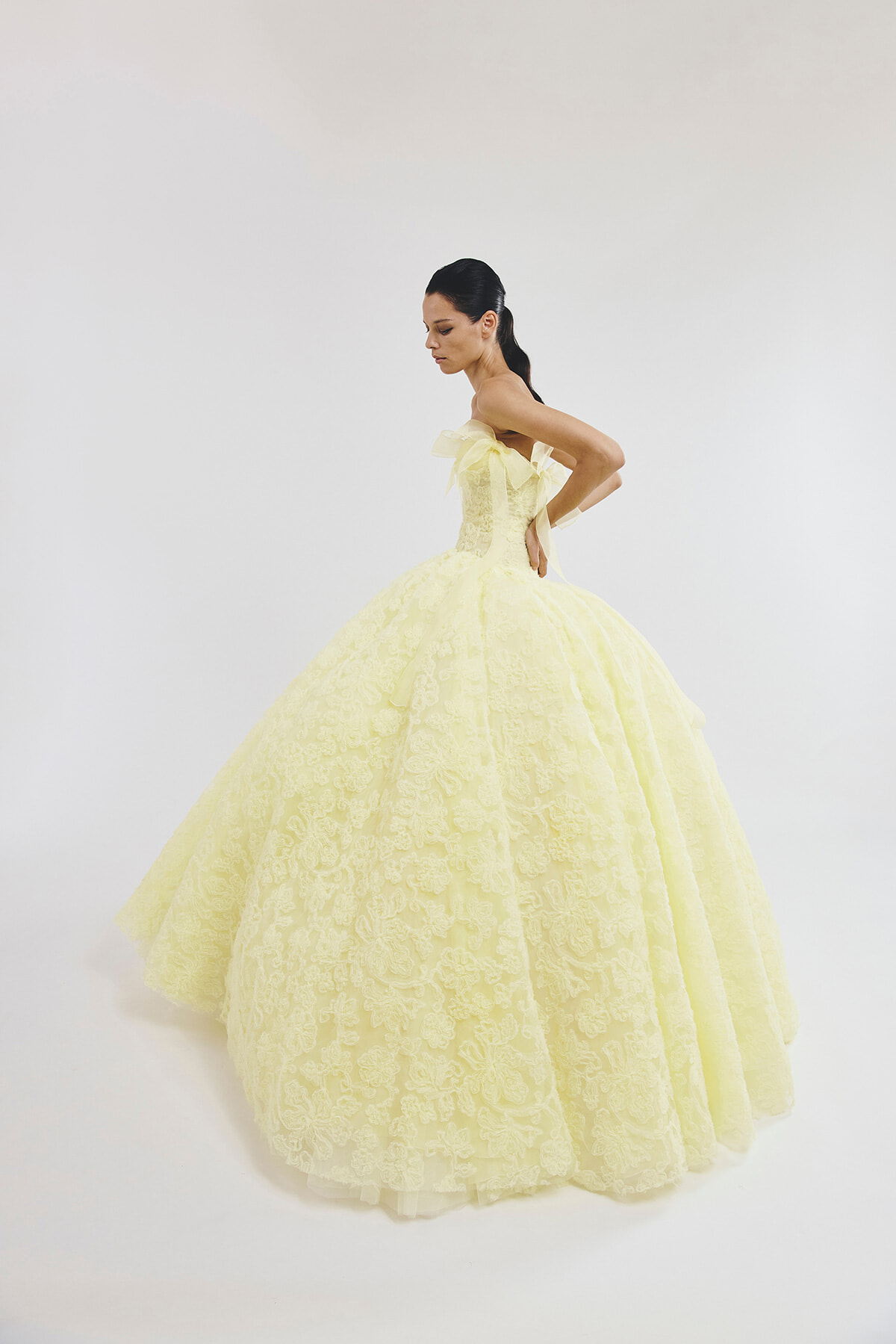 Model in a pale-yellow lace ball gown with a full, layered skirt, shown in profile for a fashion shoot.