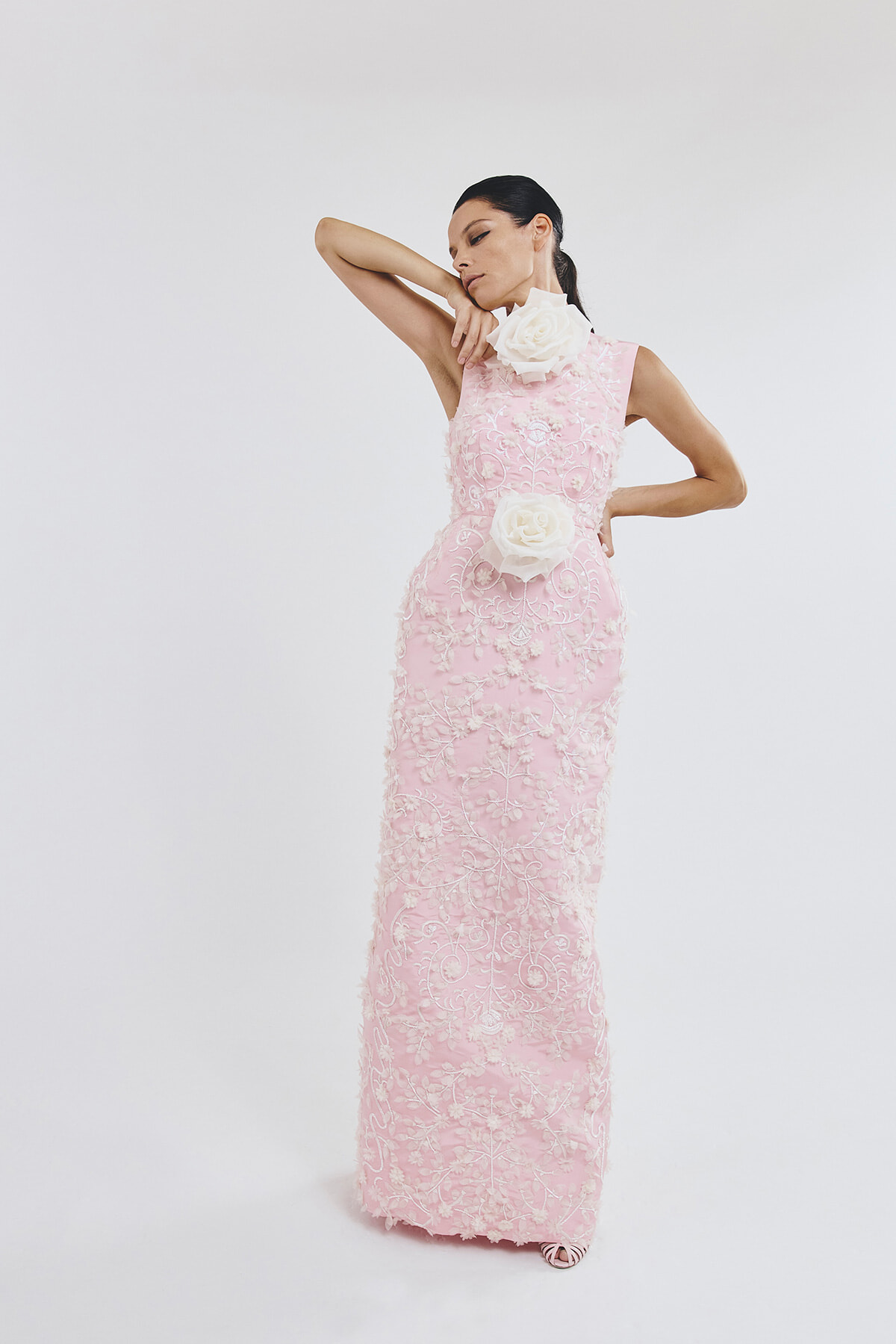 Model wearing a pink lace to-the-floor gown with white floral appliqués, posing with one arm raised and a large white rose at the shoulder and waist.