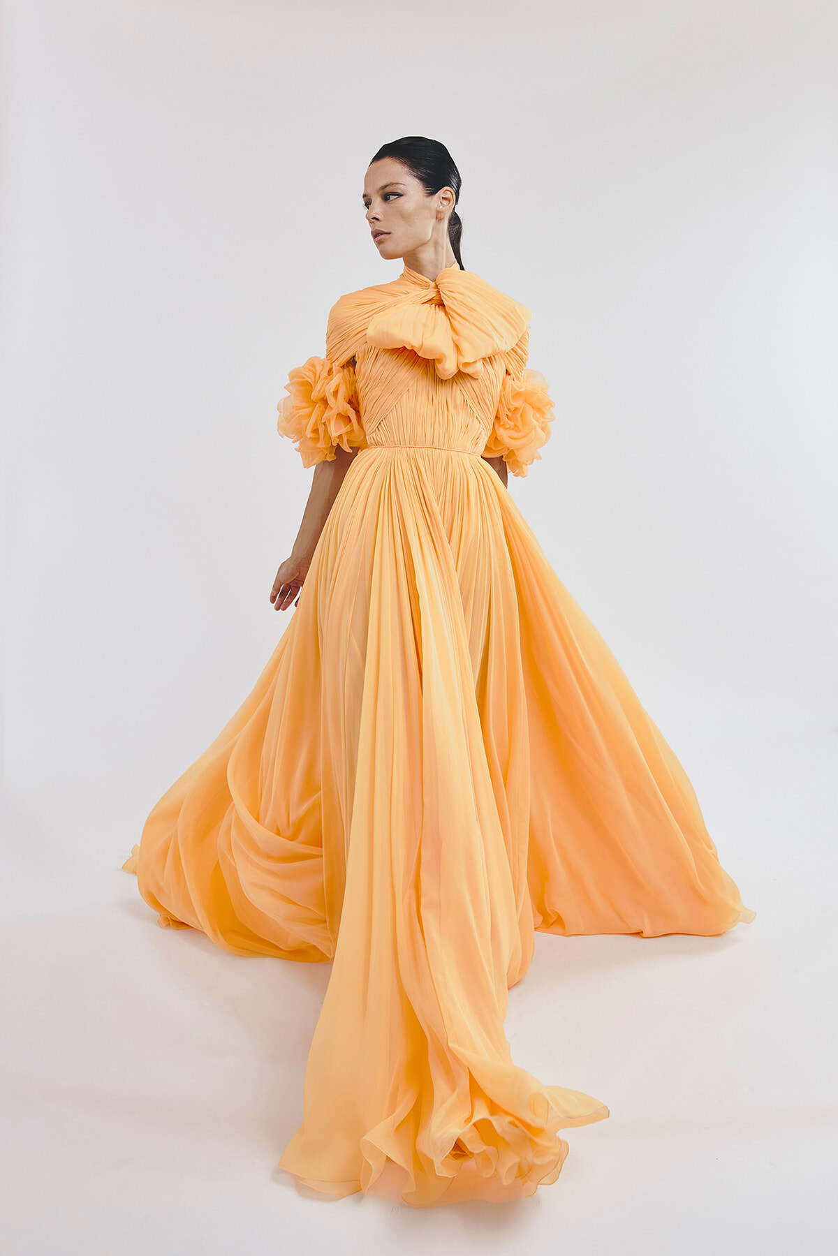 Woman in a peach floor-length gown with dramatic puff sleeves and a large bow at the neck, posing against a light backdrop.