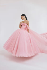 Woman in a voluminous pink ball gown with floral bodice posing gracefully with her hand near her collarbone, dress trailing behind her