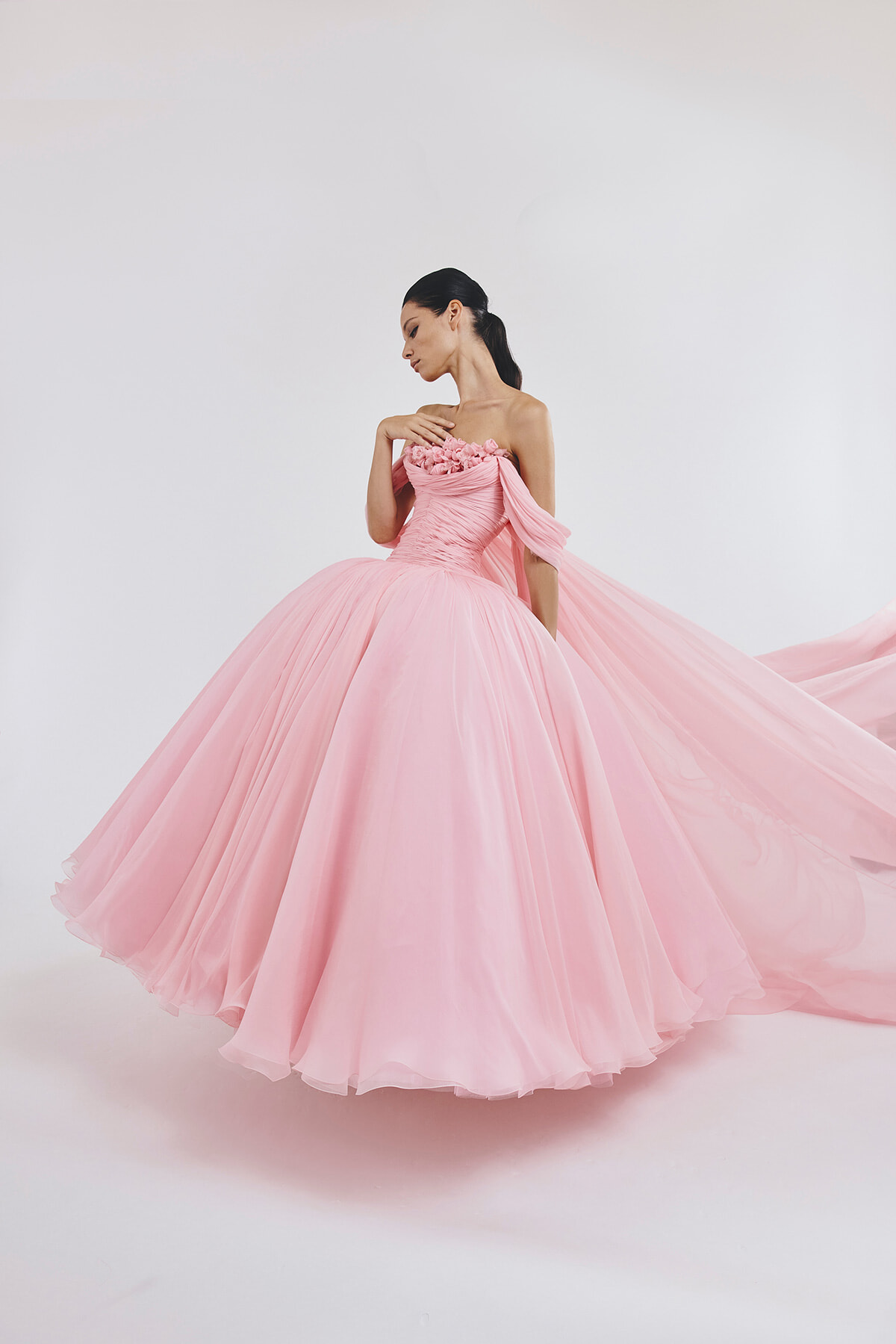 Woman in a voluminous pink ball gown with floral bodice posing gracefully with her hand near her collarbone, dress trailing behind her