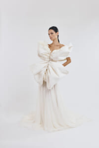 Model wearing an avant-garde white bridal gown with a large sculpted bow at the waist, off-the-shoulder sleeves, and a long flowing train in a white studio setting