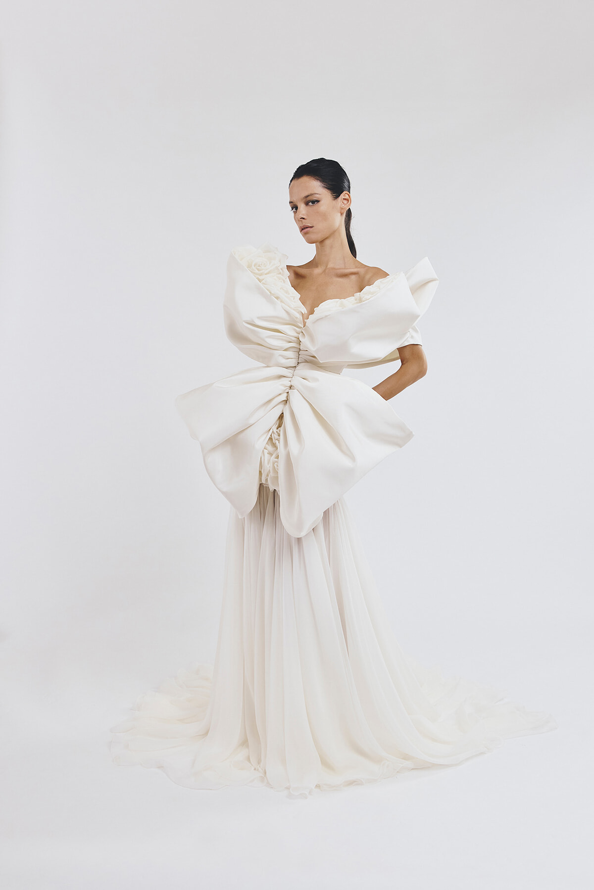 Model wearing an avant-garde white bridal gown with a large sculpted bow at the waist, off-the-shoulder sleeves, and a long flowing train in a white studio setting