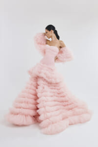 Model in a strapless pink ball gown with voluminous ruffled skirt and fluffy sleeves, posing against a white backdrop.