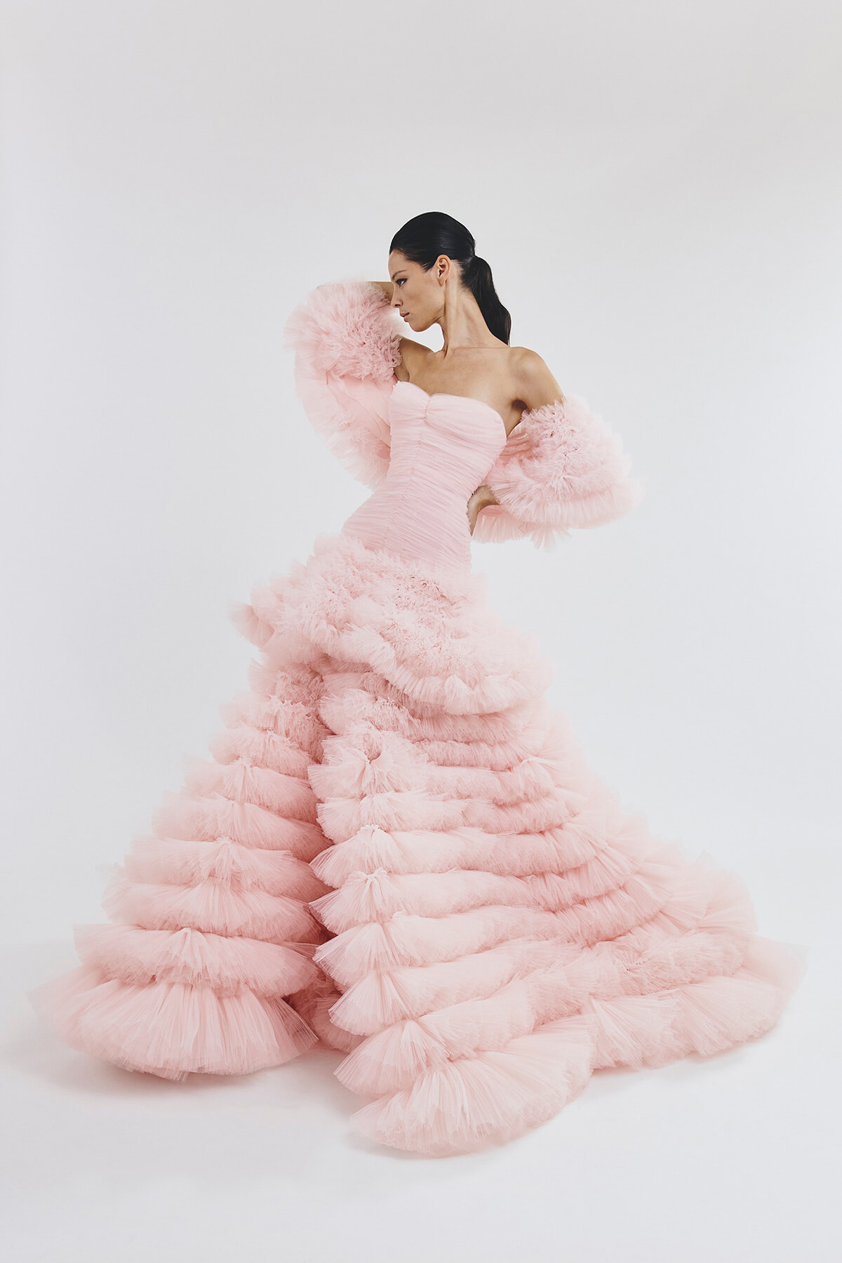Model in a strapless pink ball gown with voluminous ruffled skirt and fluffy sleeves, posing against a white backdrop.