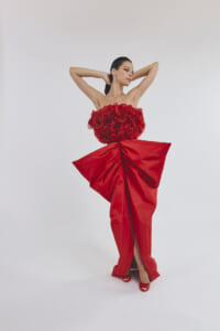 Model wearing an avant-garde red gown with a rose-bedecked bodice and sculpted skirt, posing with arms raised.