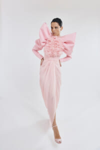 Model in a pale pink gown with oversized bow shoulders and a floral ruffle bodice, standing with hands on hips against a white studio backdrop, wearing pink heeled sandals.