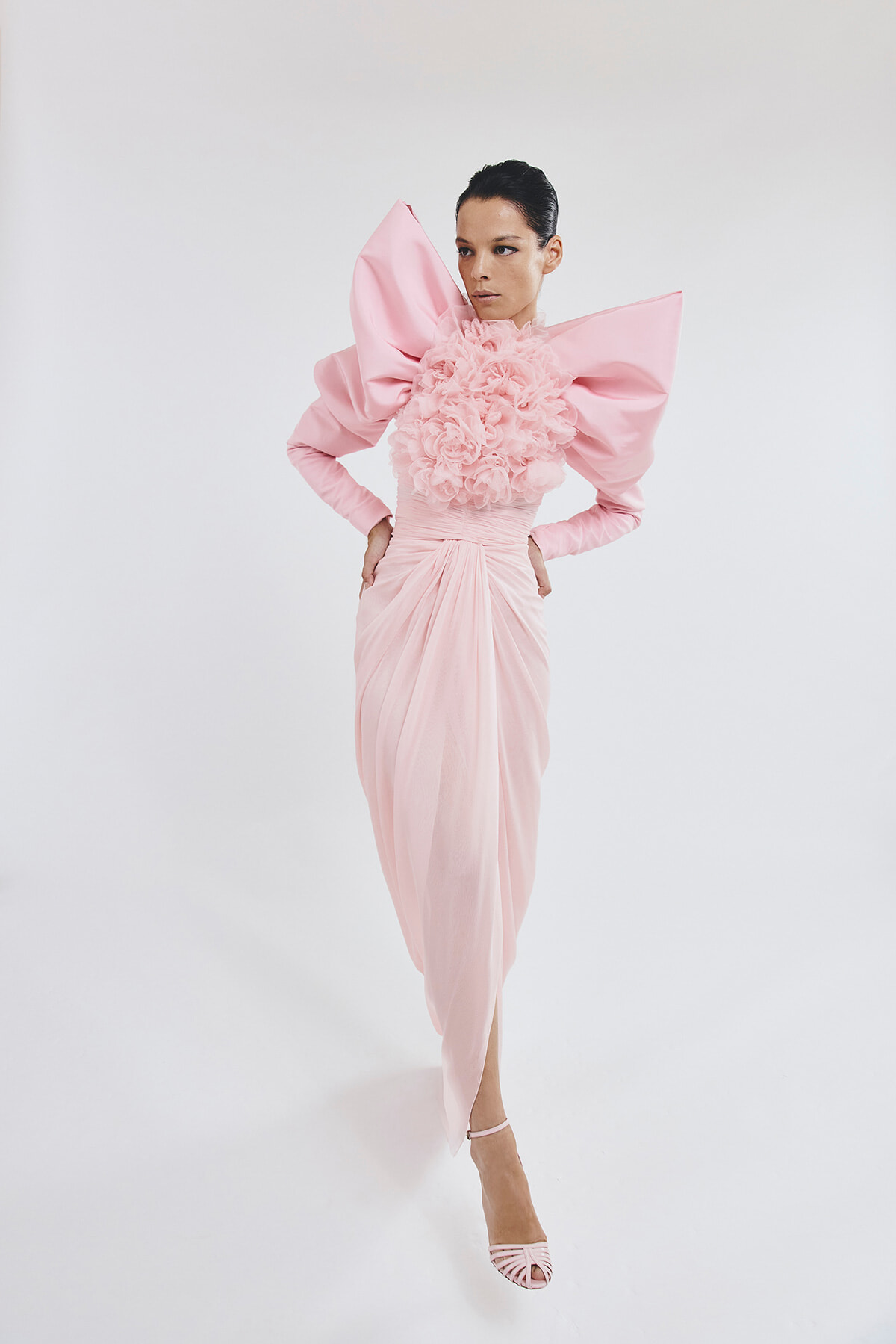 Model in a pale pink gown with oversized bow shoulders and a floral ruffle bodice, standing with hands on hips against a white studio backdrop, wearing pink heeled sandals.