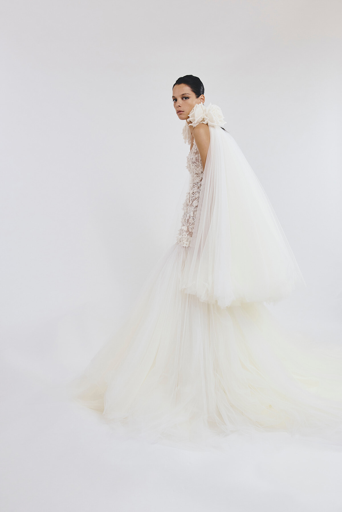 Model wearing an ivory wedding gown with a lace bodice and a voluminous sheer tulle cape/skirt, posed against a pale background.
